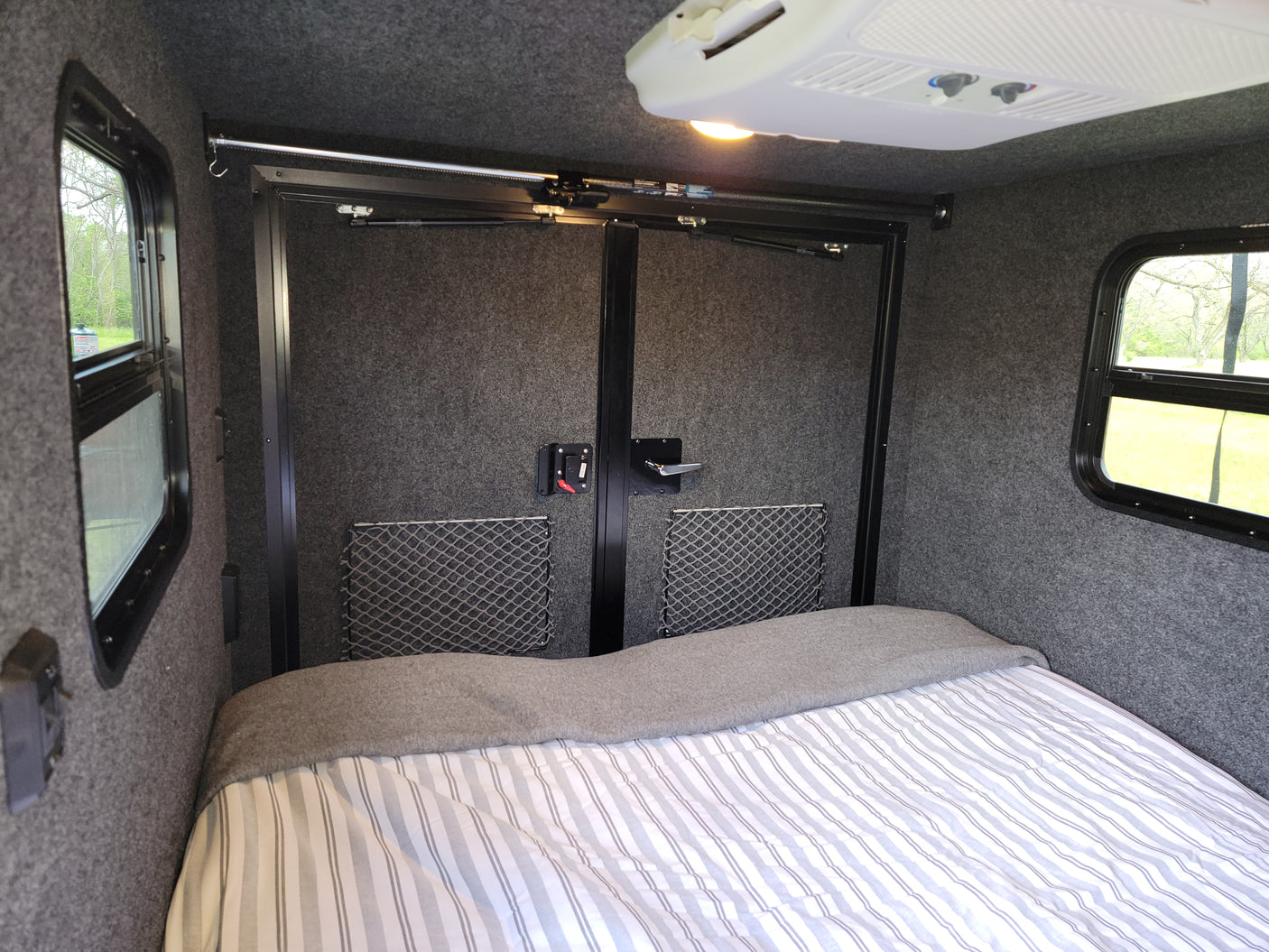 Cozy RV rental interior with bed, striped bedding, windows, storage nets, and overhead AC.
