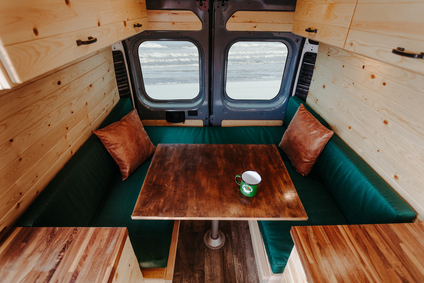 An inviting RV rental interior with wood details, comfy green seating, and a table overlooking a serene beach scene.