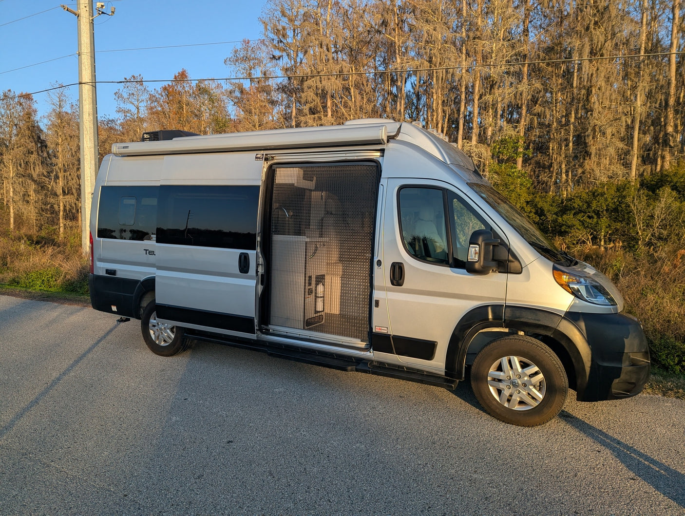 Silver rv rental camper van with open sliding screen door, parked on a road beside mossy trees on a sunny day.