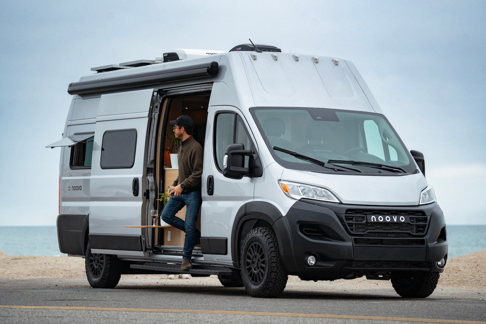 A man exits a modern NOOVO camper van RV rental, parked on a road next to an ocean beach under an overcast sky.