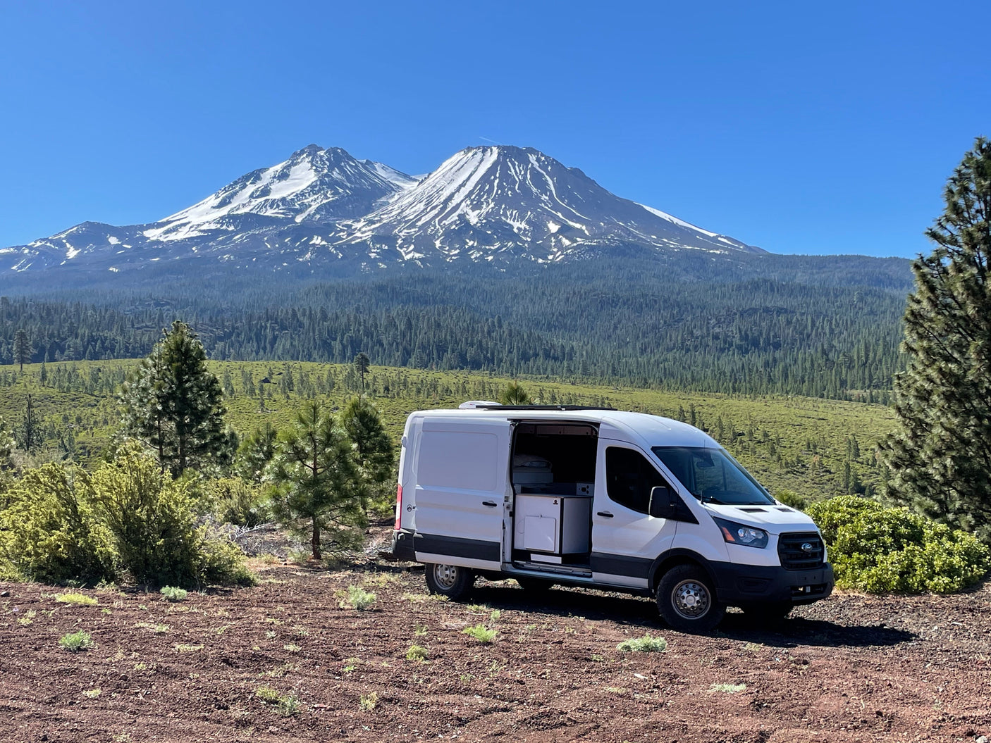 A white Ford Transit RV rental van, door open, sits on dirt, facing snow-capped Mount Shasta & forested valley under blue sky.