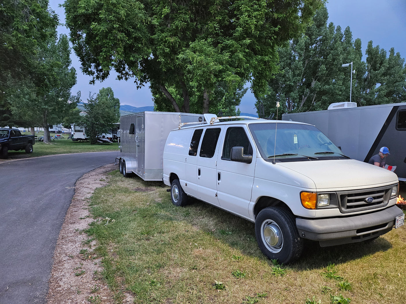 White Ford van towing a silver cargo trailer, parked at a spacious RV campground. Perfect for an RV rental journey.