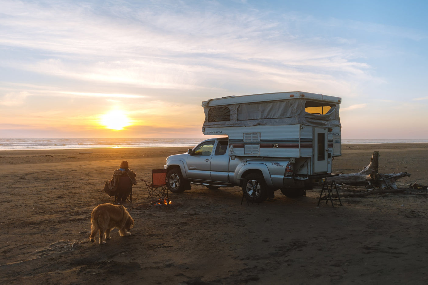 Truck camper RV rental parked on a sandy beach at sunset. A person and dog relax by a crackling campfire near the ocean.