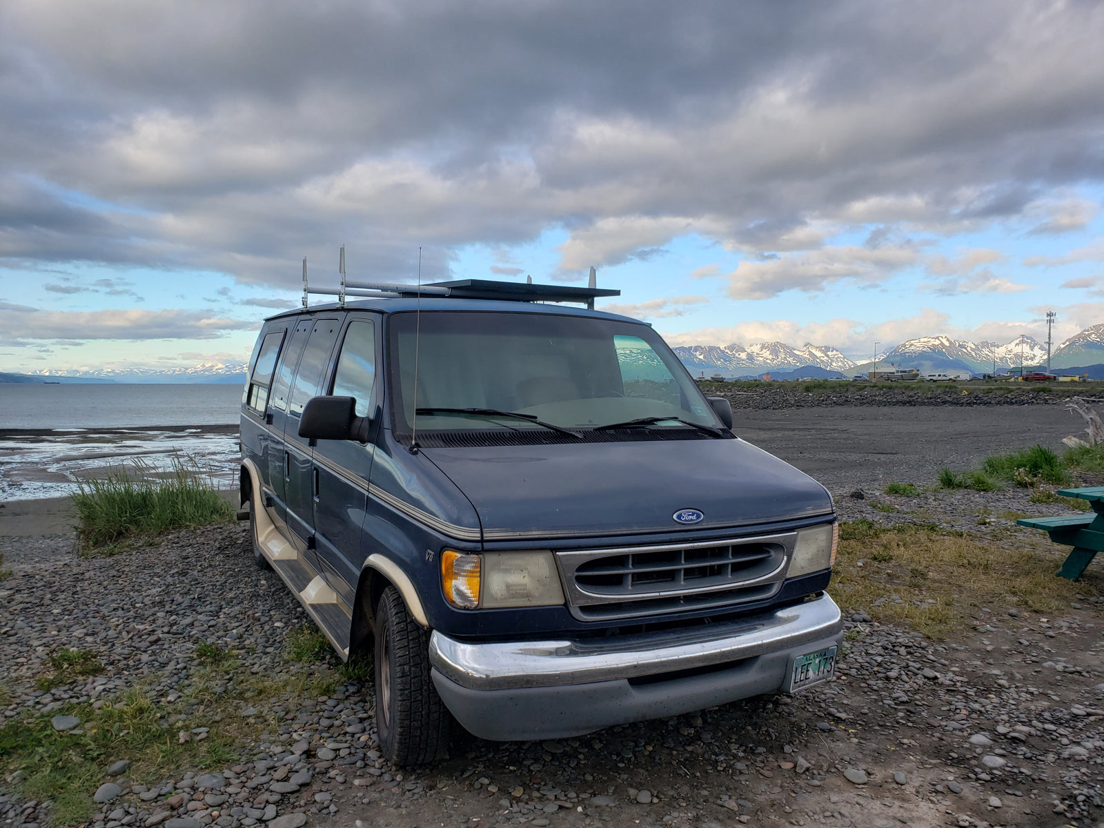 A blue Ford van, perfect for an RV rental adventure, parked on an Alaskan rocky beach facing snow-capped mountains.