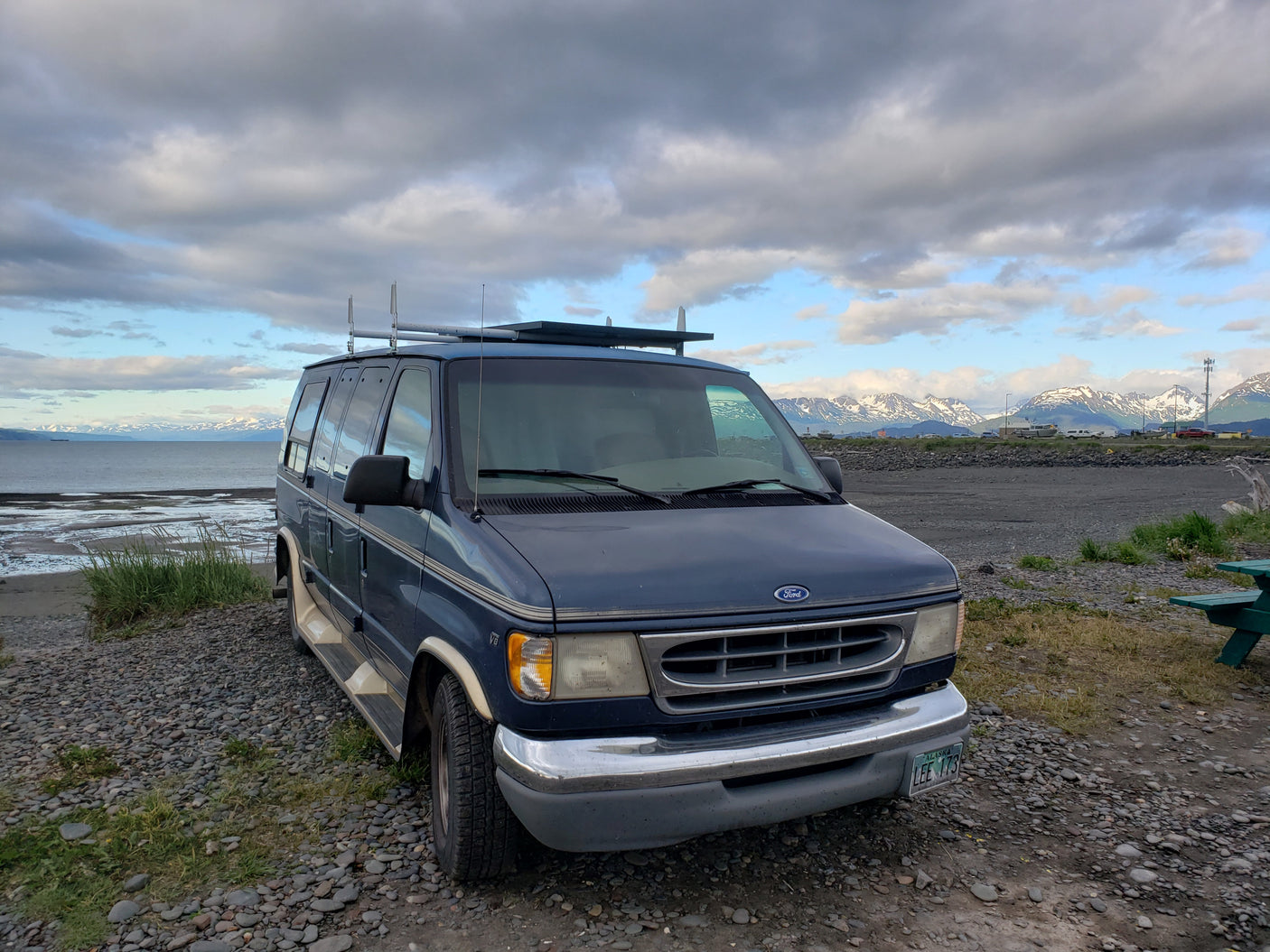 A blue Ford van, perfect for an RV rental adventure, parked on an Alaskan rocky beach facing snow-capped mountains.
