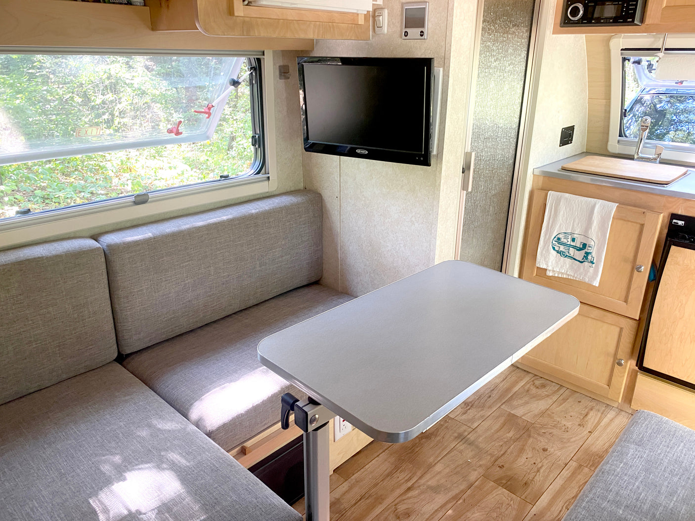 Inside a compact rv rental: dinette, small kitchen, and TV. Bright window view of nature.