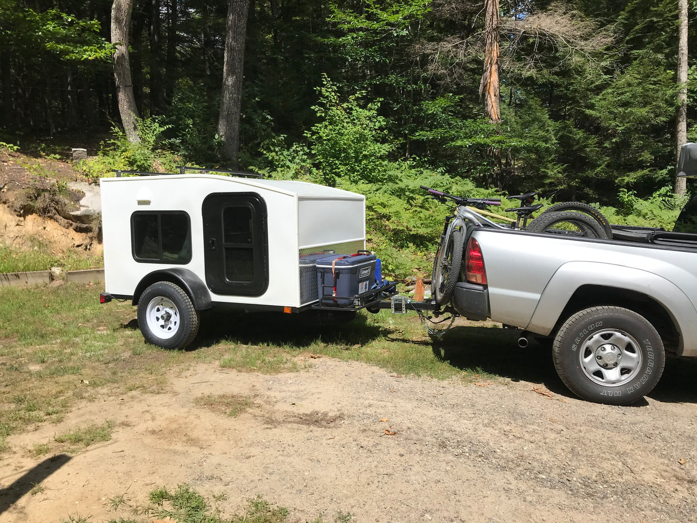 A white teardrop trailer (like an RV rental) is hitched to a silver pickup truck with mountain bikes, parked in a wooded area.