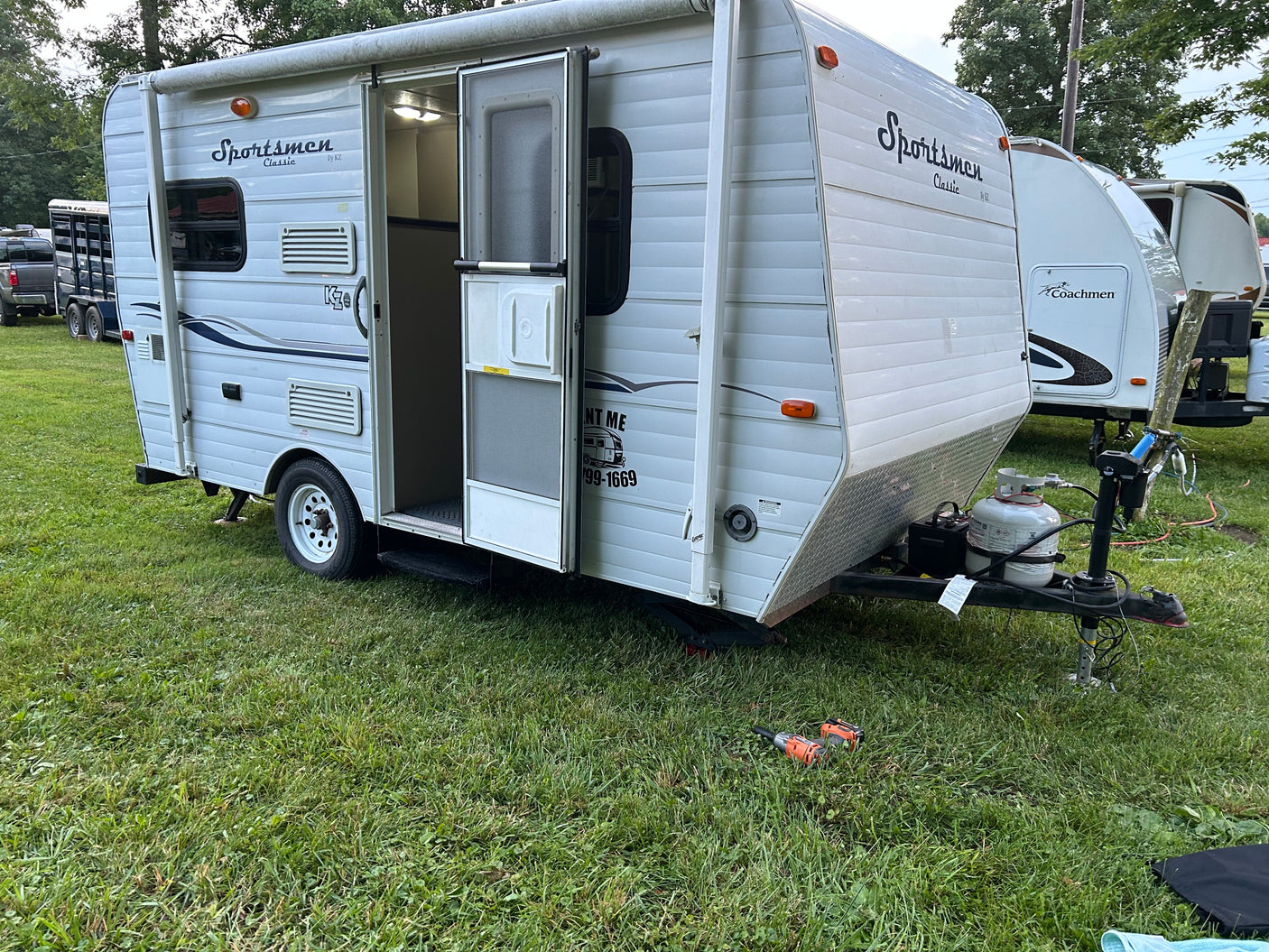 This white Sportsmen RV rental features an open door, propane tanks, and is parked on grass.