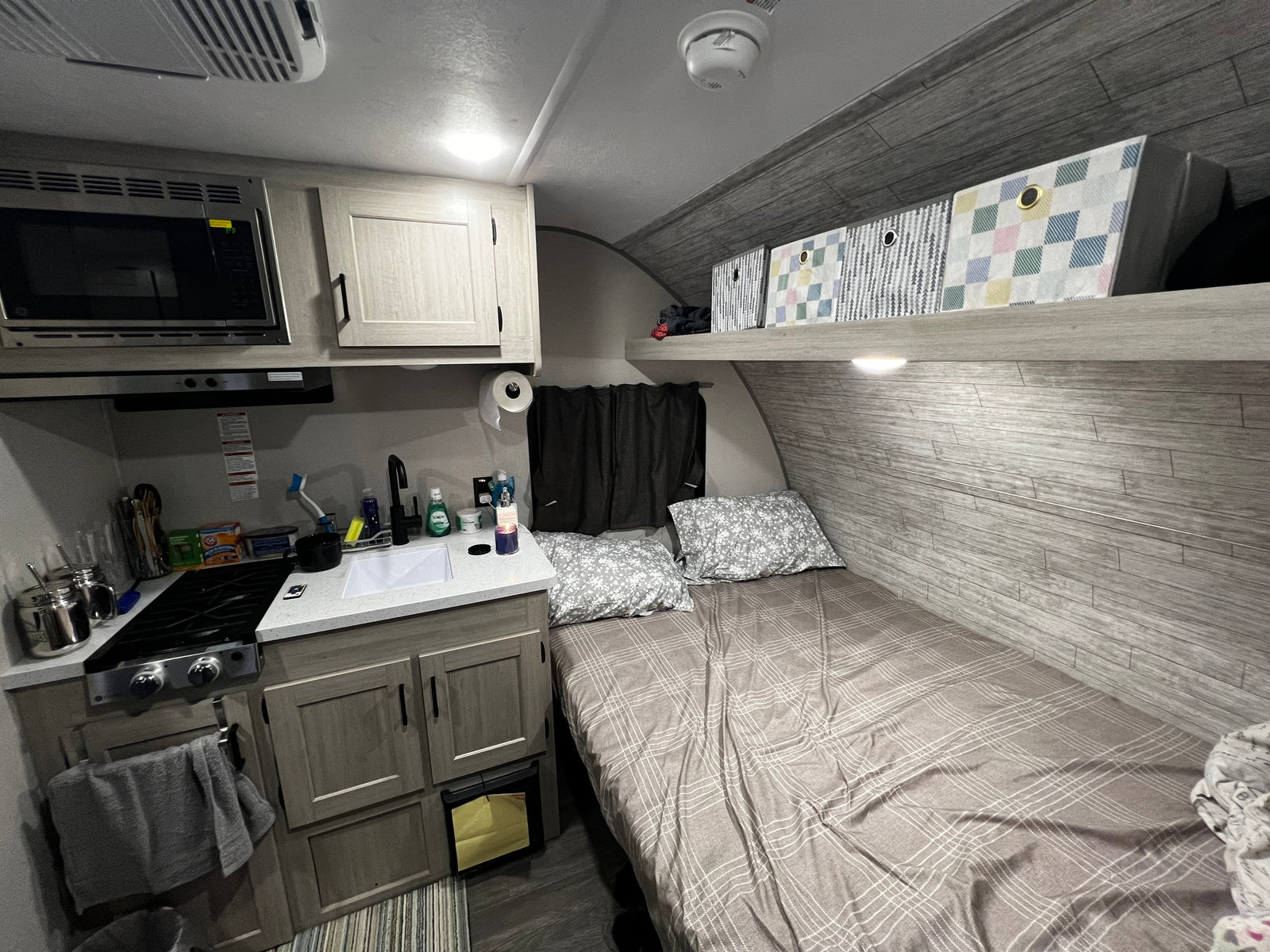 RV rental interior: compact kitchen (microwave, stove, sink), cozy bed with pillows, and overhead storage in neutral tones.