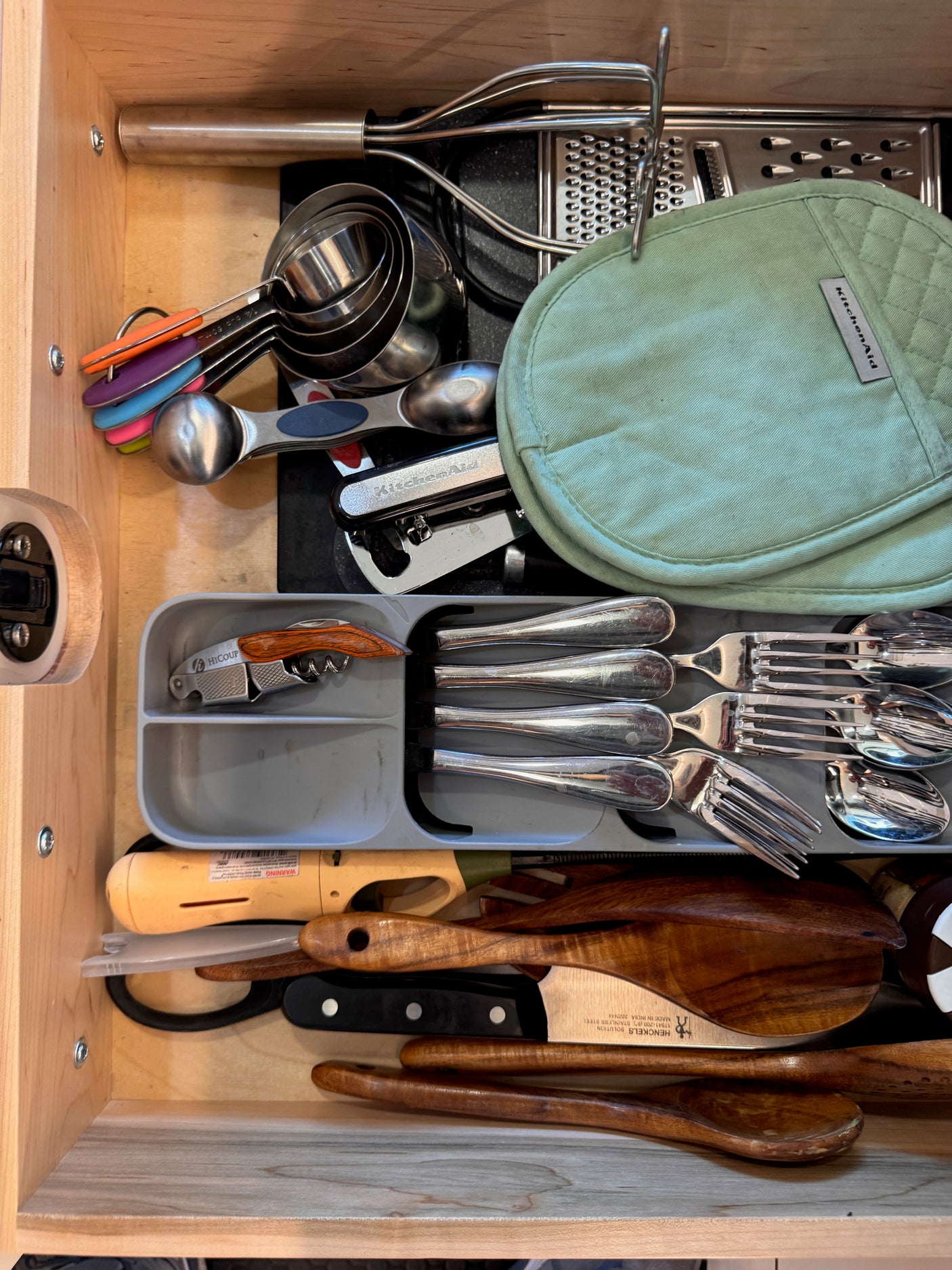 Organized rv rental kitchen drawer: measuring spoons, grater, cutlery, KitchenAid mitt, and wooden utensils.