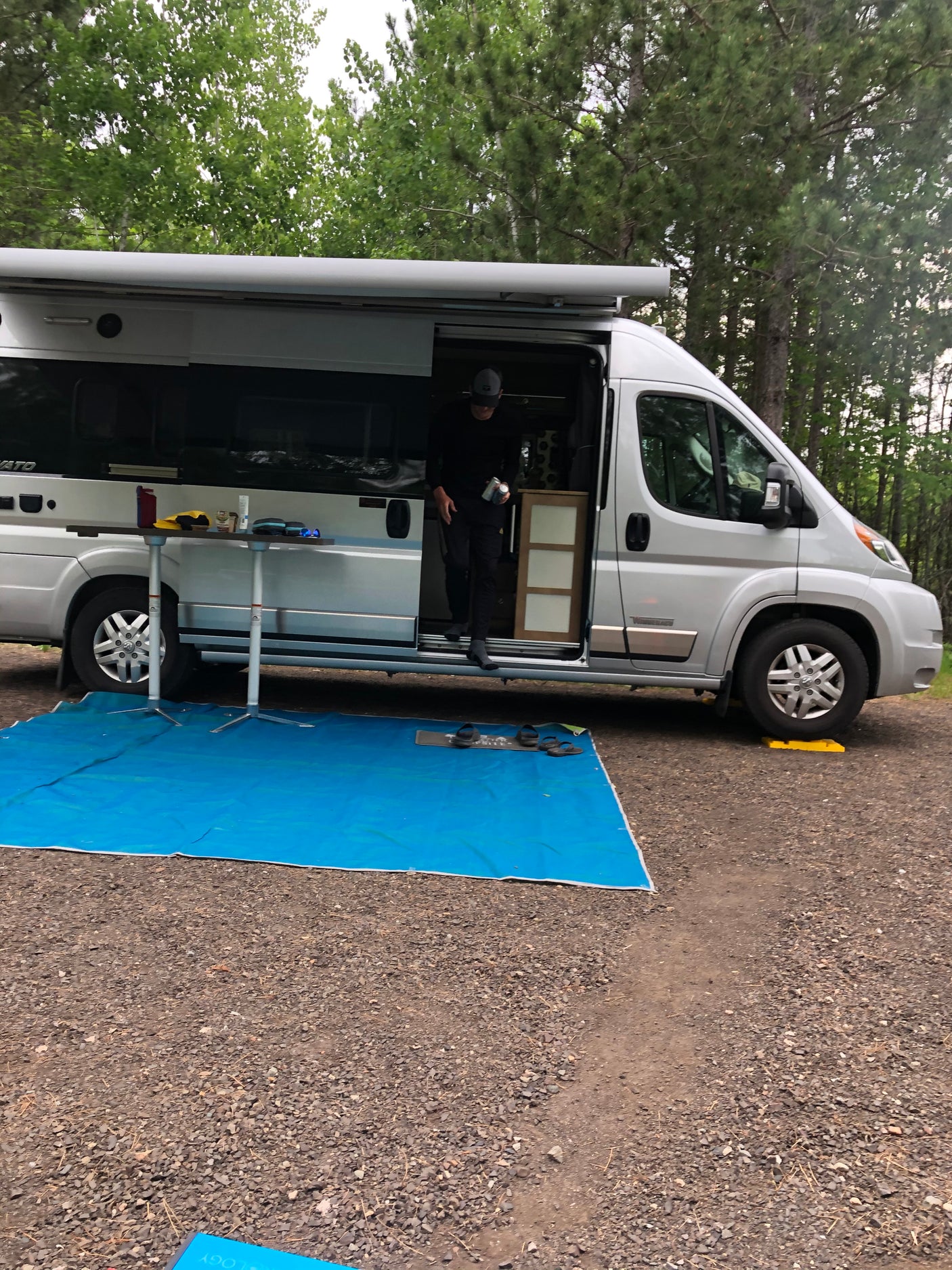 Person steps from a silver RV rental van at a gravel campsite. Outdoor table and blue tarp create a cozy setup.