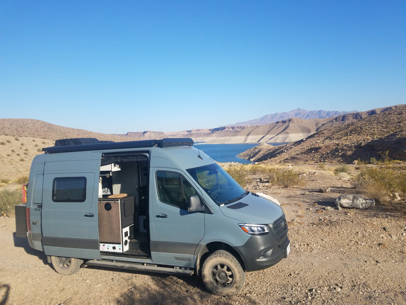 This grey RV rental is parked with its side door open, revealing its interior by a beautiful blue lake and arid mountains.