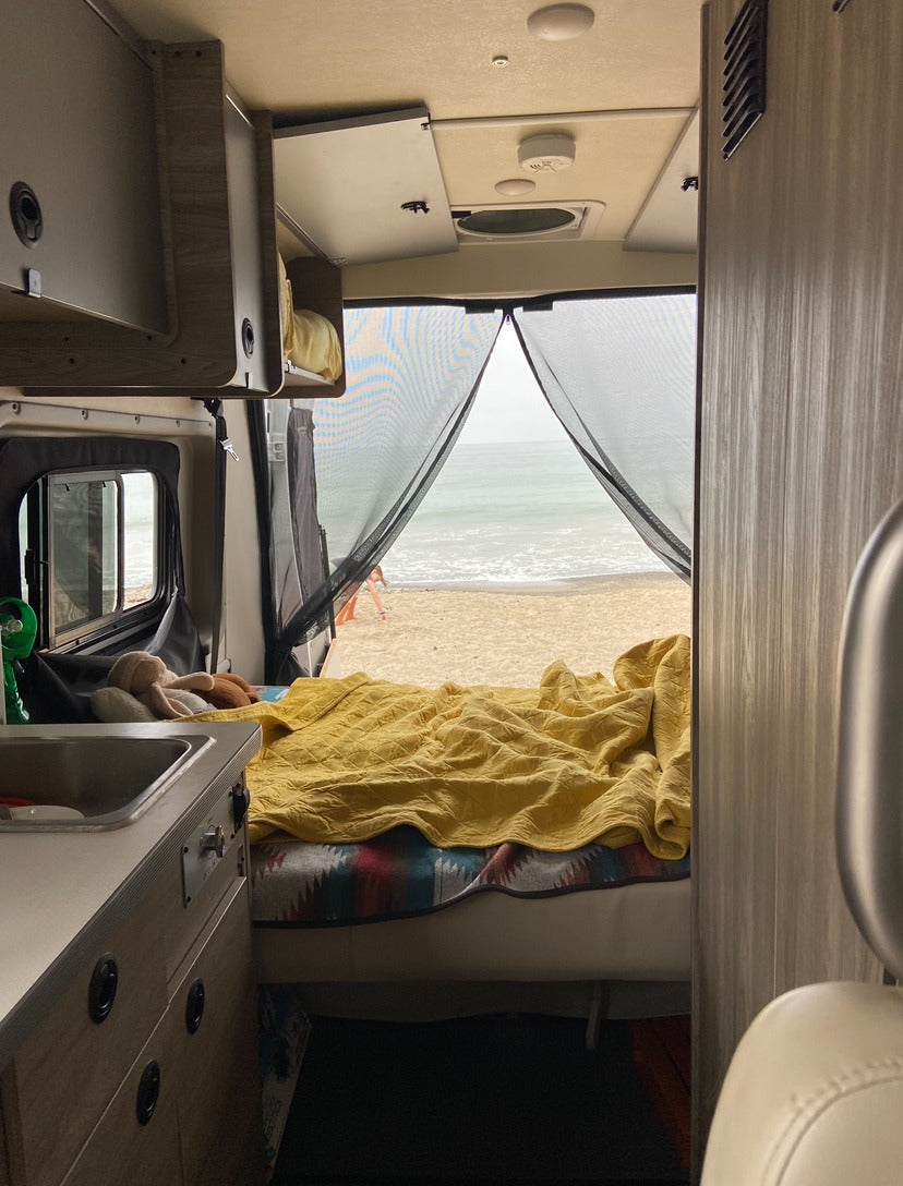 Inside an RV rental, a comfy bed and kitchenette face a stunning beach and ocean view, seen through a mesh screen.