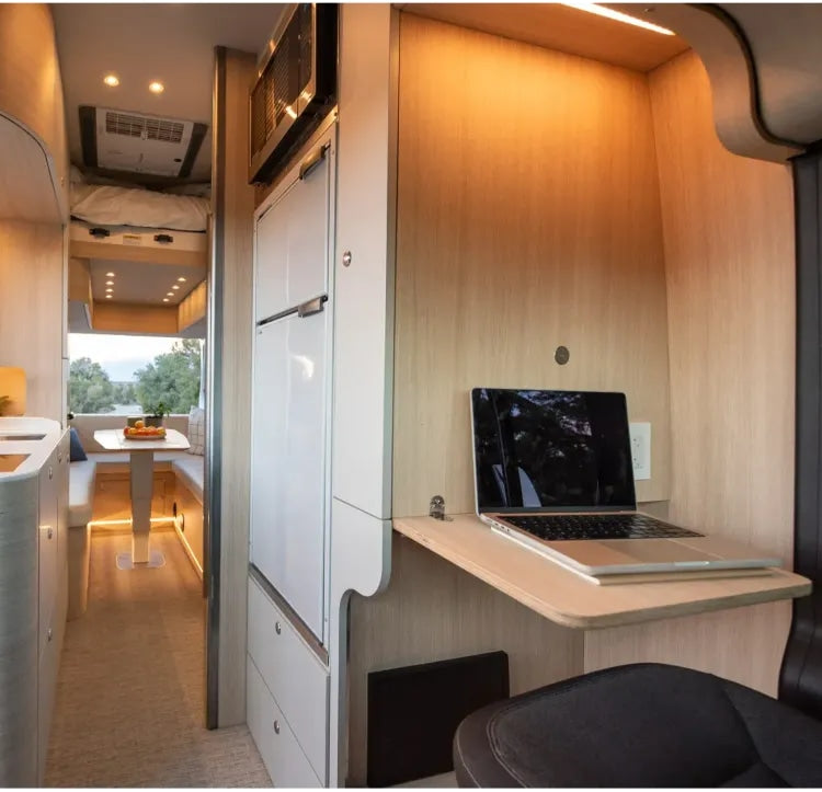Modern RV rental interior features a laptop on a fold-down desk, a compact kitchen, and a dining area.