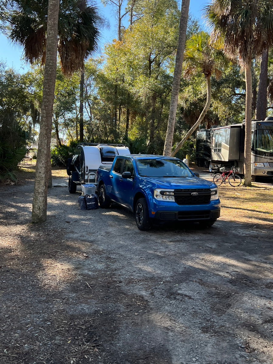 Blue truck with rv rental camper at a sunny campsite. Larger motorhomes and palm trees in this serene camping setting.