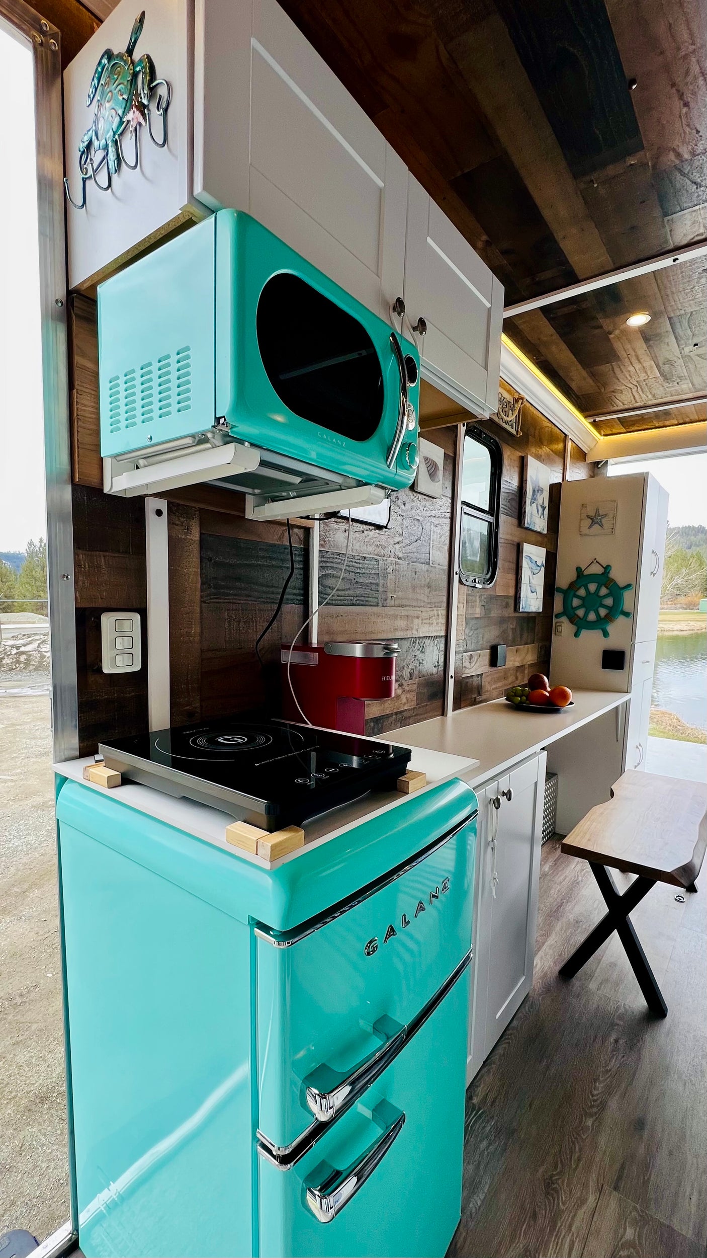 This compact RV rental kitchen boasts a retro teal Galanz fridge, microwave, and induction cooktop, offering scenic pond views.