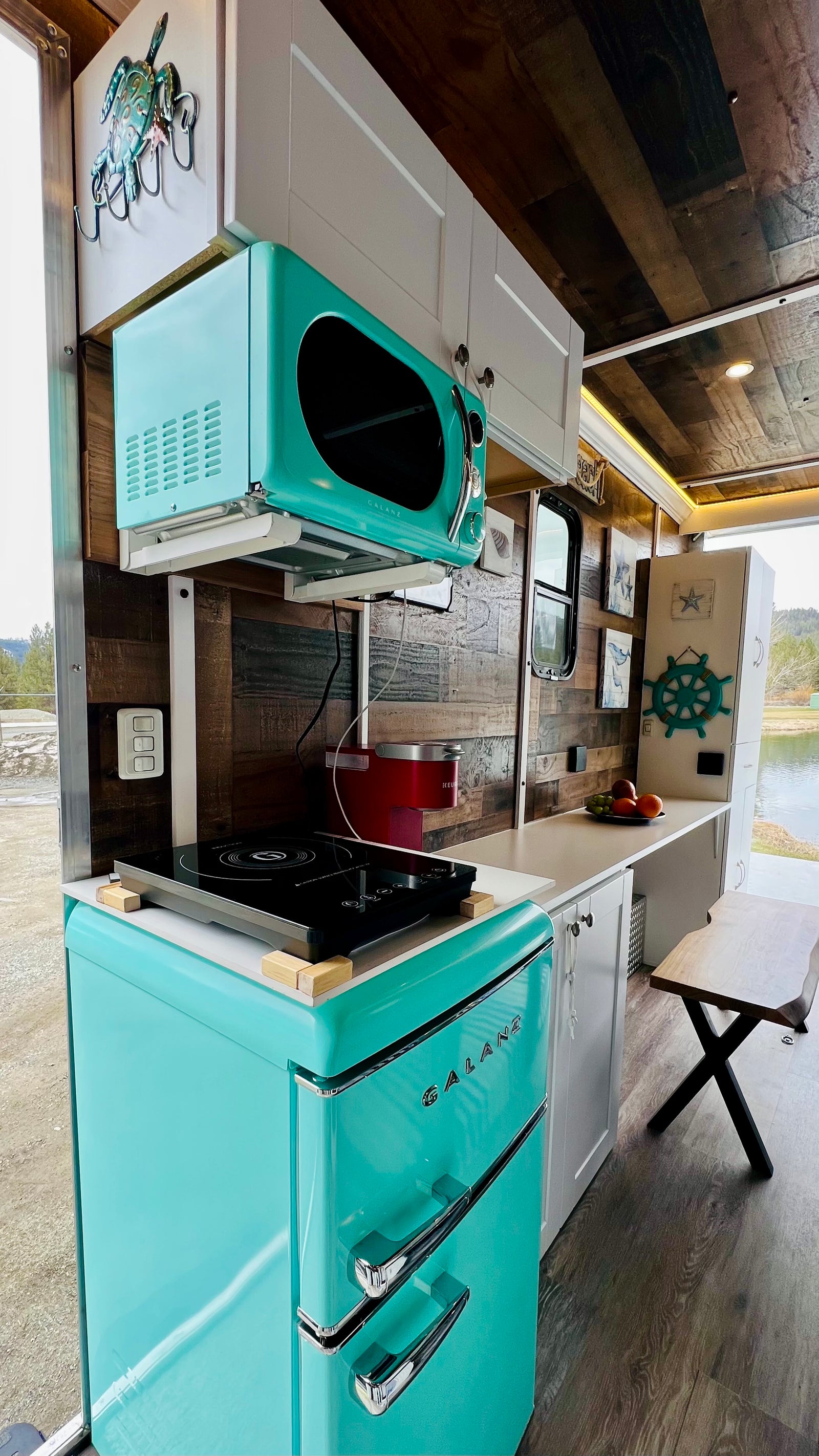 This compact RV rental kitchen boasts a retro teal Galanz fridge, microwave, and induction cooktop, offering scenic pond views.