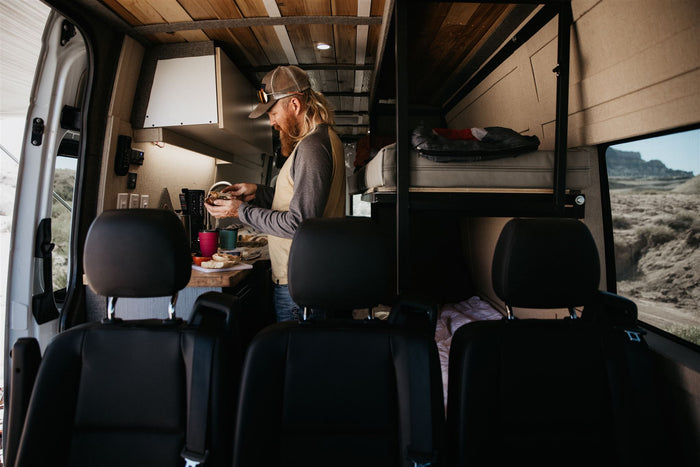 RV rental interior: A man preparing food in a cozy camper van kitchen with bunk beds & desert views.