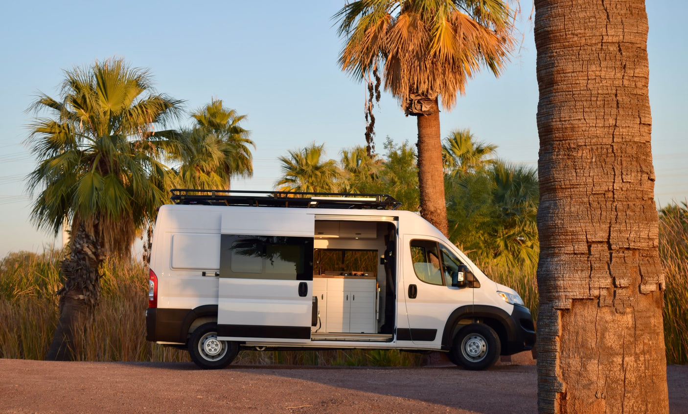 White camper van RV rental parked on a dirt path with its side door open, revealing a modern kitchen. Palm trees surround it.
