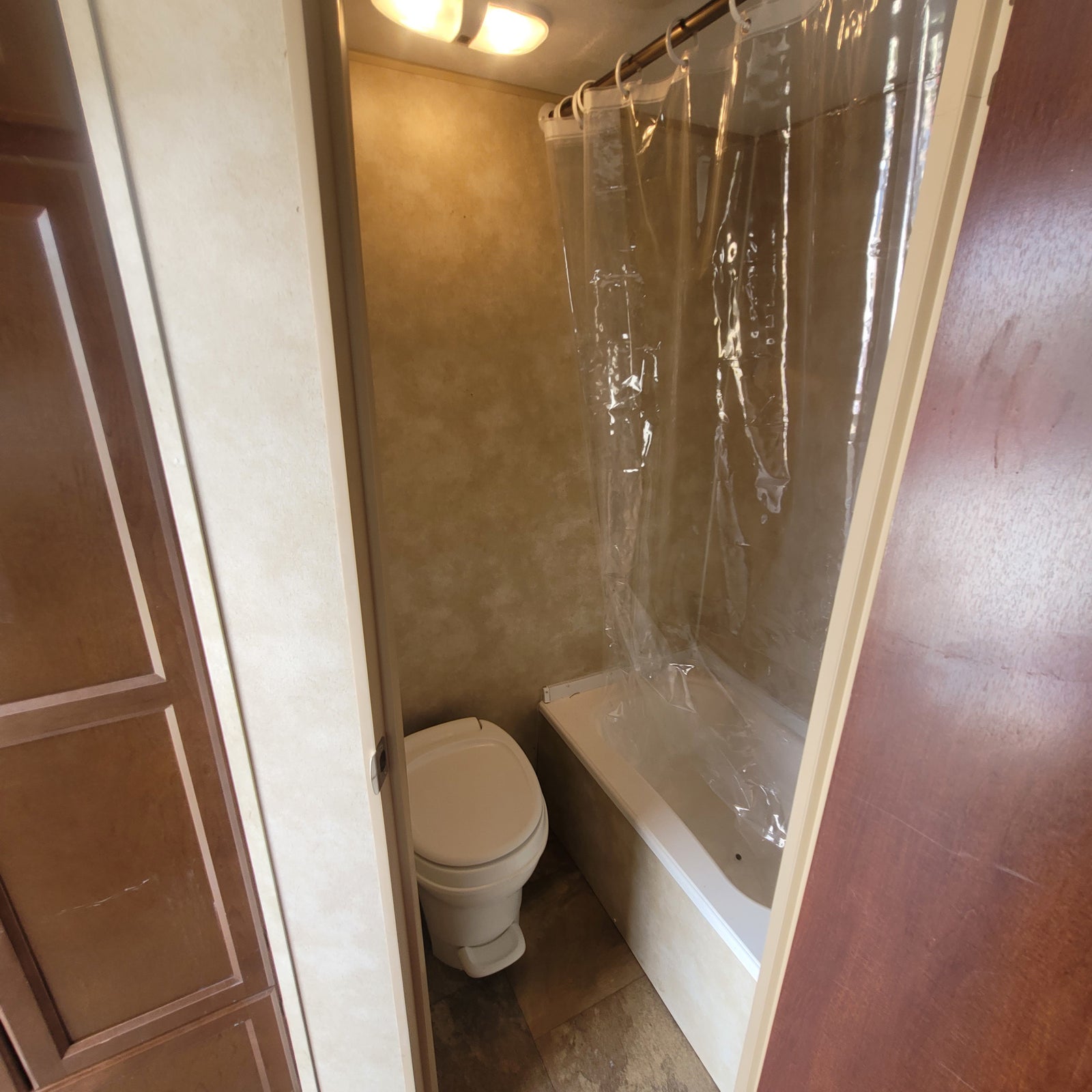 RV rental bathroom with a toilet on the left and a bathtub/shower with a clear curtain on the right, lit by an overhead fixture.