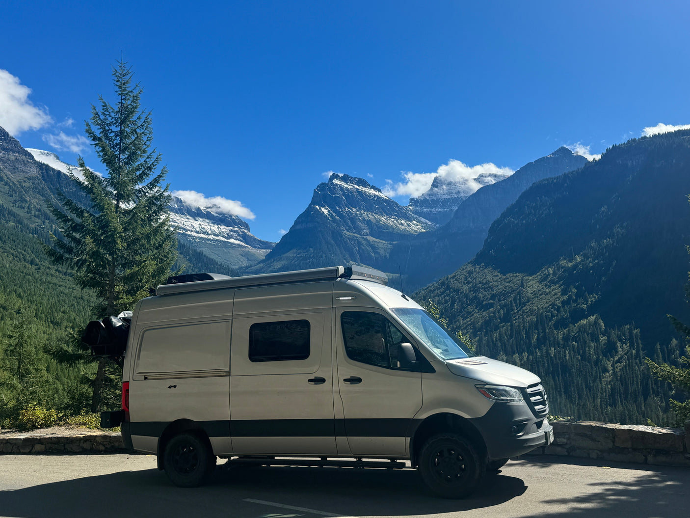 Explore nature's beauty! A light-colored RV rental is parked at a scenic mountain overlook, surrounded by snow-capped peaks and tall trees.