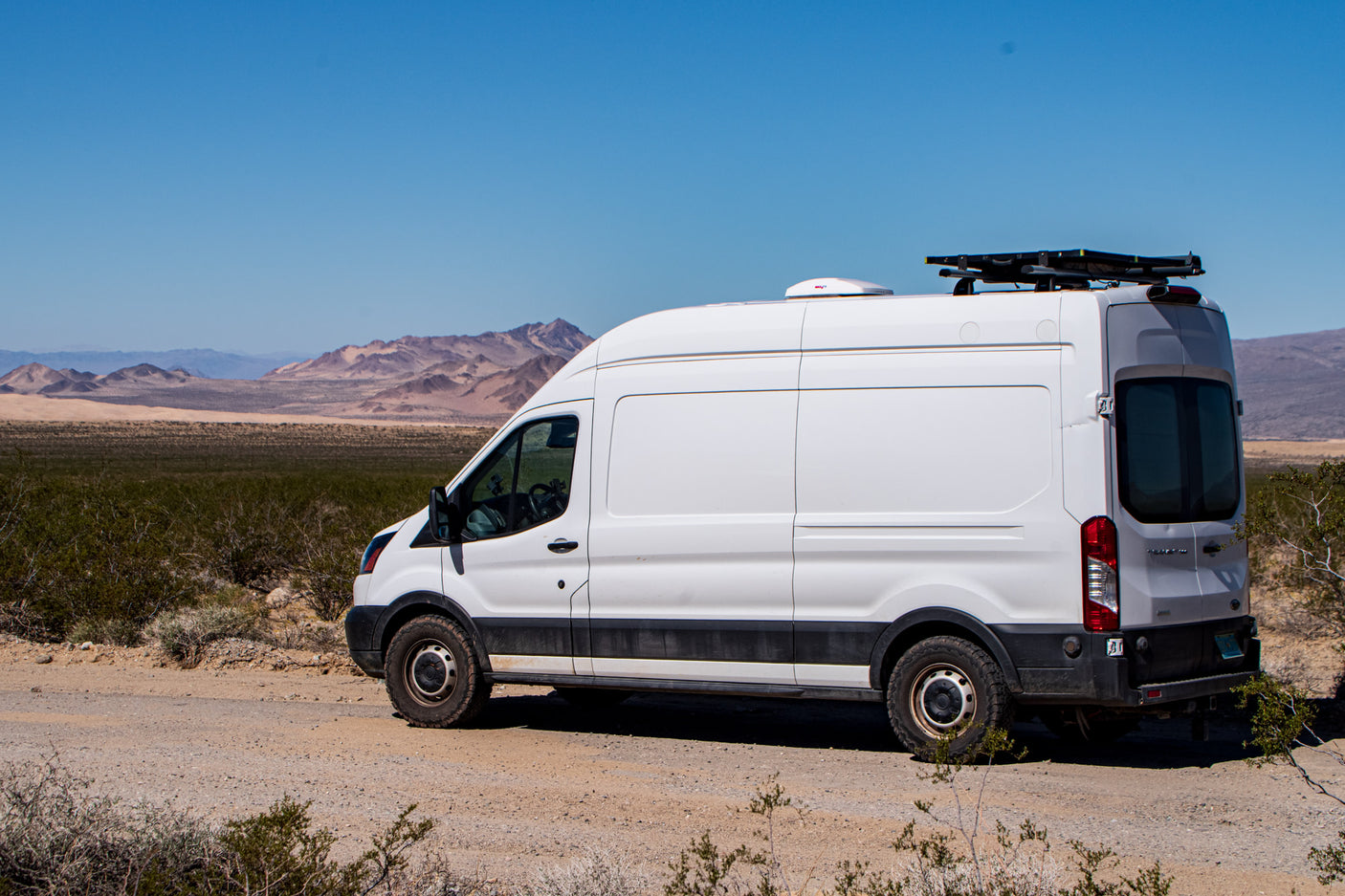 White high-roof RV rental van, off-road ready, parked on a desert dirt path with mountains under a blue sky.
