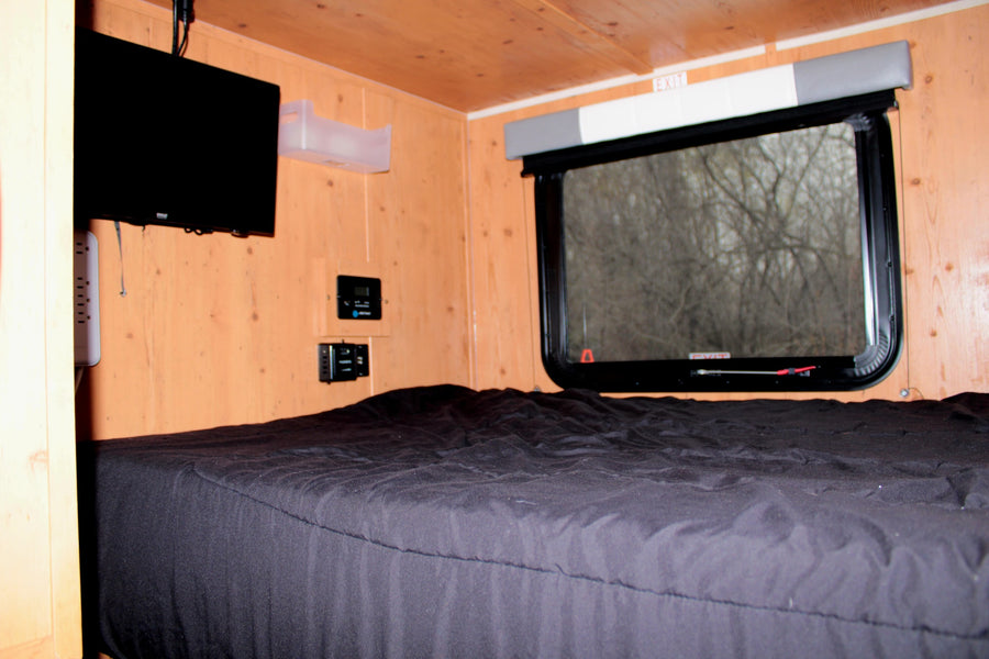 RV rental bedroom: black bed, mounted TV, wood walls, window with blind, controls, & EXIT sign.