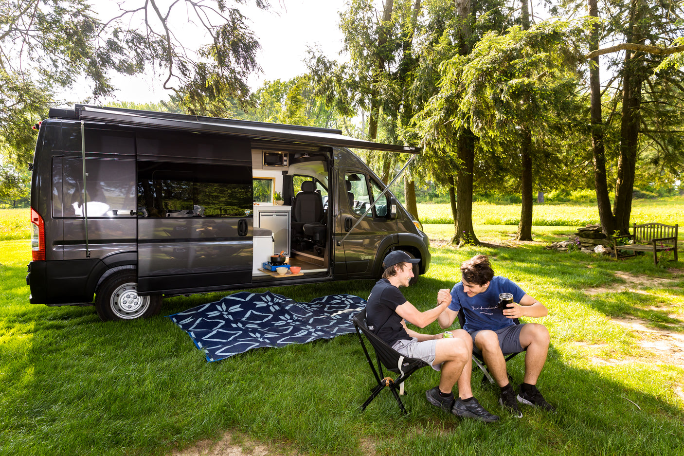 Friends enjoy their RV rental adventure. A camper van is parked with an awning extended, two men relax in camping chairs outside.