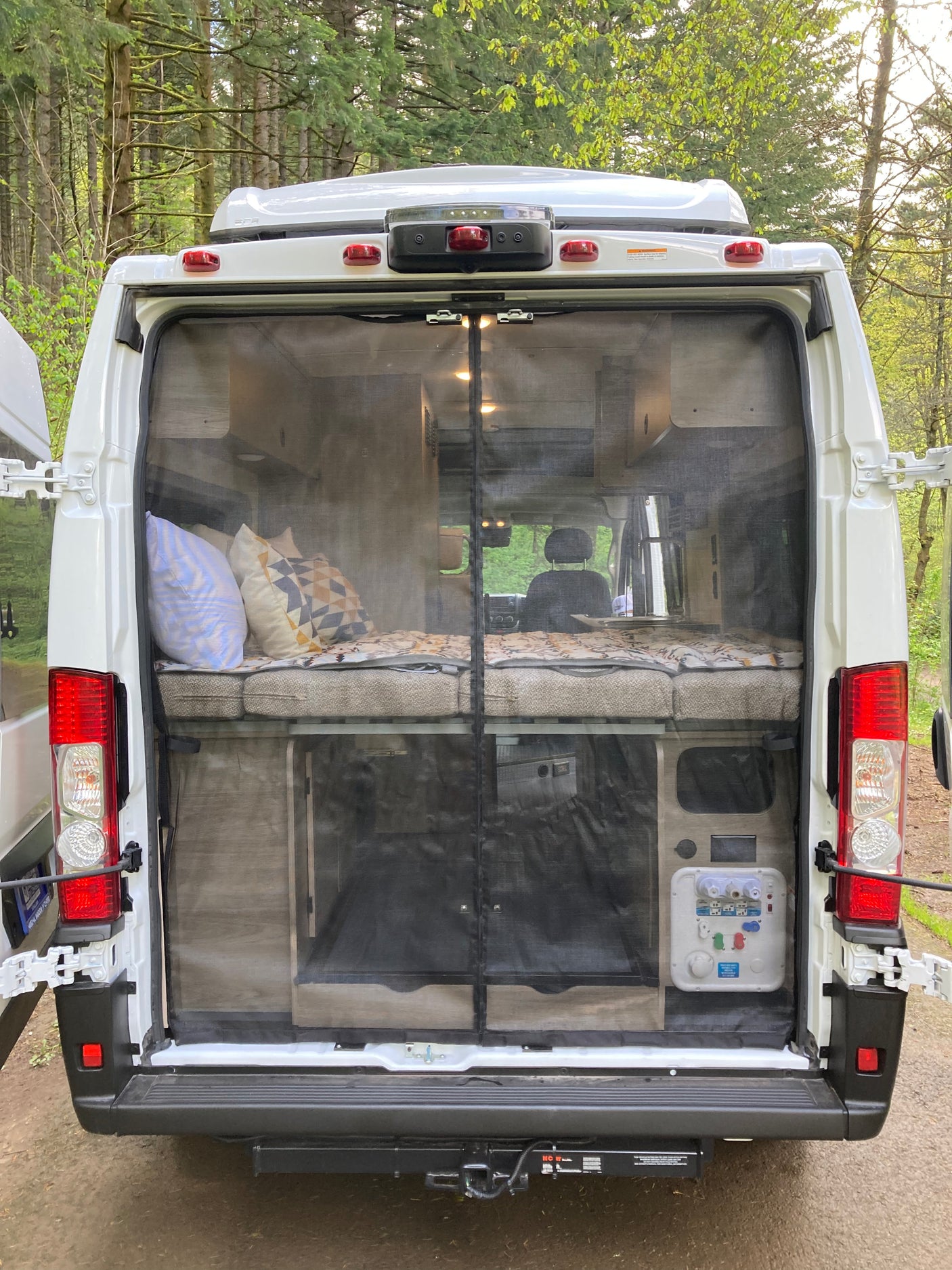 Open back of an rv rental camper van, revealing a bed, storage, and utility panel behind a protective bug screen. Ideal for camping.
