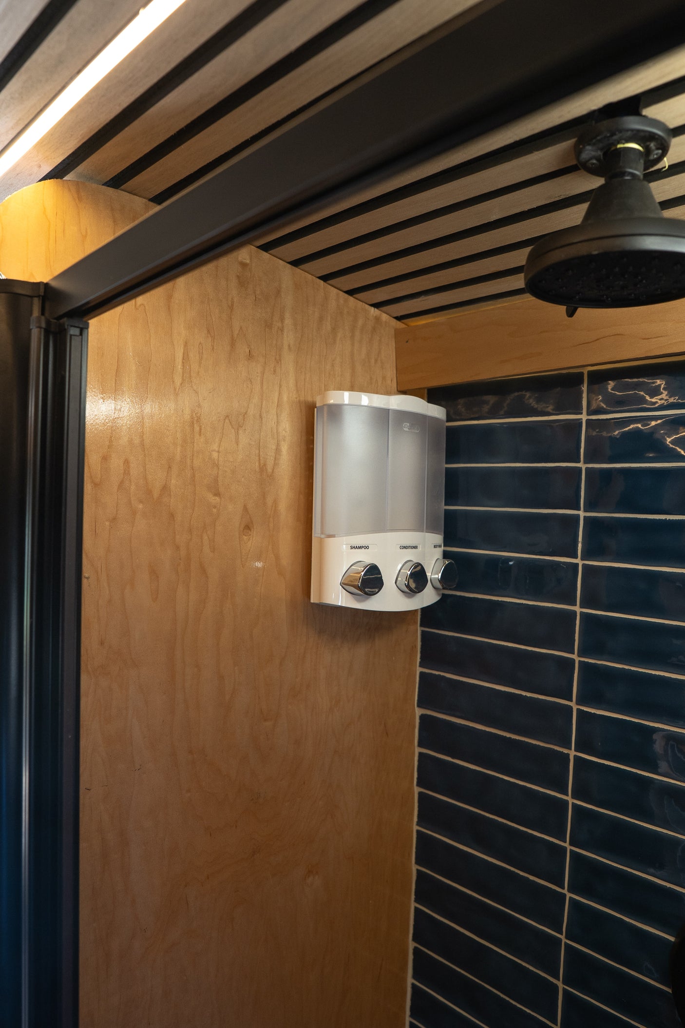 Inside an RV rental shower: warm wood panels, dark blue subway tiles, a black shower head, and a convenient soap dispenser.