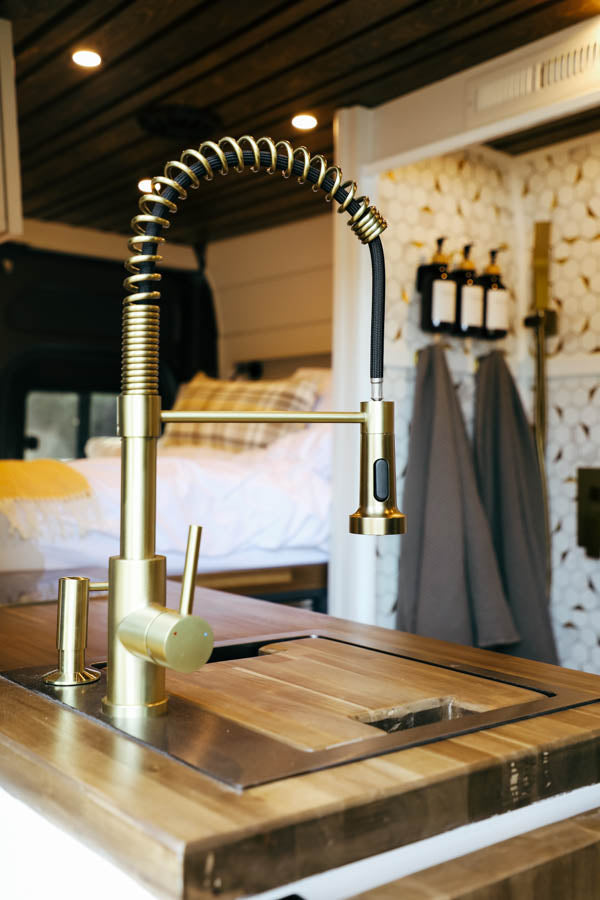 Stylish RV rental interior: close-up of a modern brass faucet, wood counter, and compact kitchen setup near the bed.