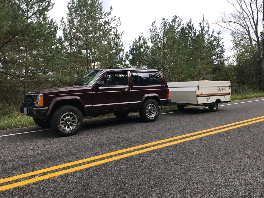 Dark red Jeep Cherokee towing a white Jayco J pop-up camper on a roadside. Great for RV rental camping!
