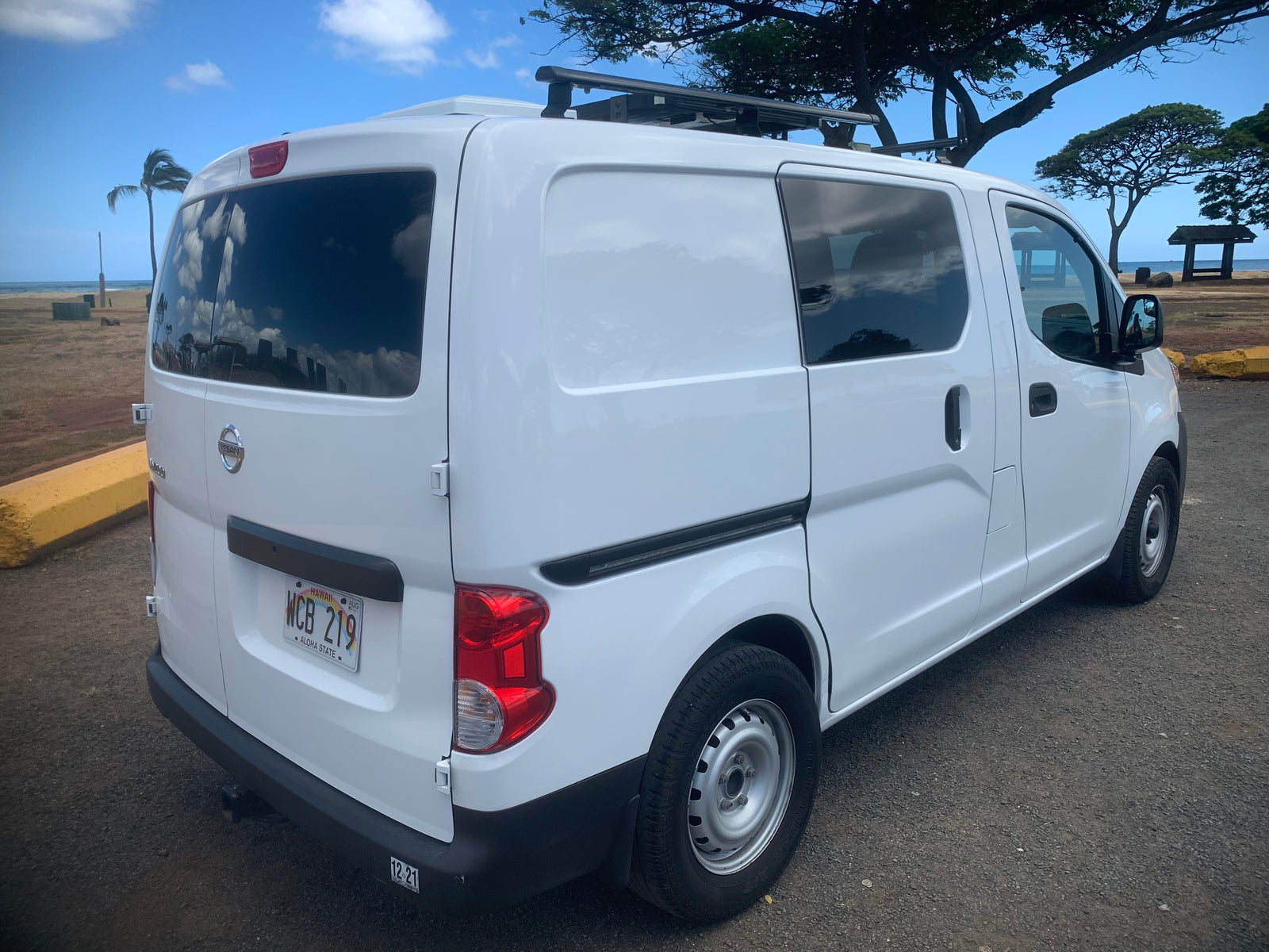 White camper van RV rental with roof rack, parked on a dirt lot next to a Hawaiian beach with palm trees.