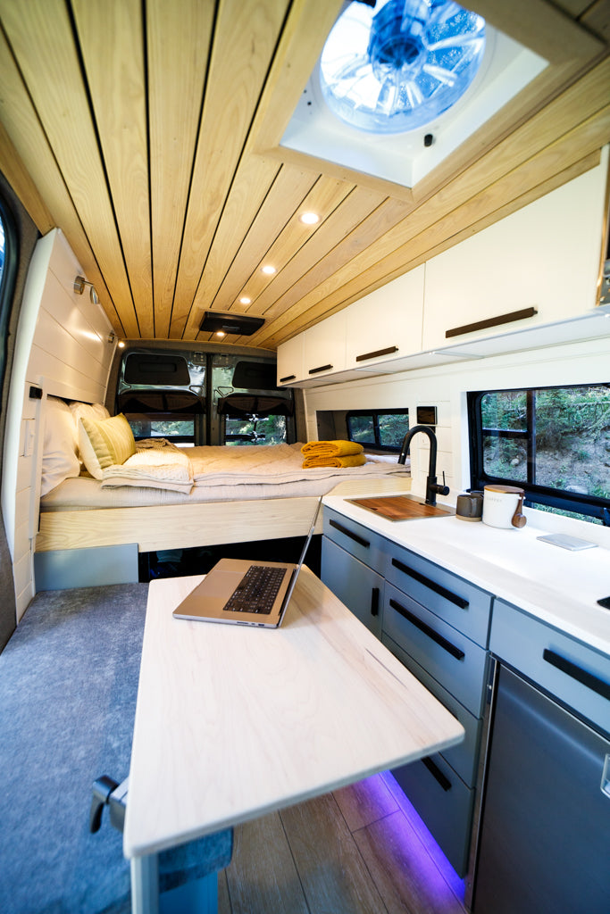 Modern RV rental interior with a cozy bed, sleek kitchen, and pull-out desk for a laptop, set against a wooden ceiling.