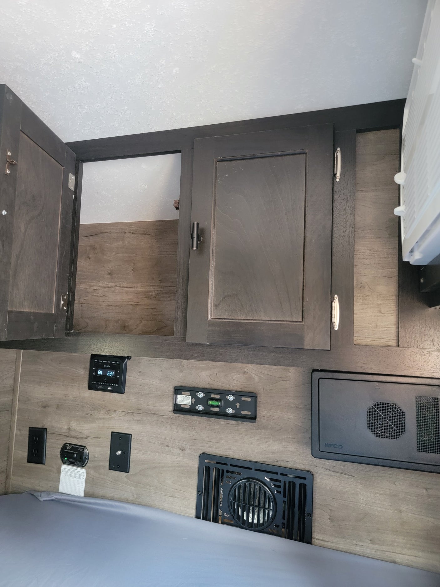 RV rental interior: wood-look cabinets (one open), bed, control panels, electrical outlets, and floor vent.