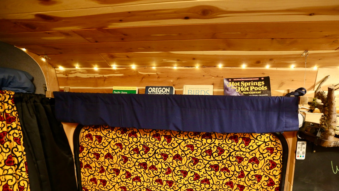 Inside a comfortable RV rental: wooden ceiling, string lights, a shelf of travel books, and a colorful patterned bed area.