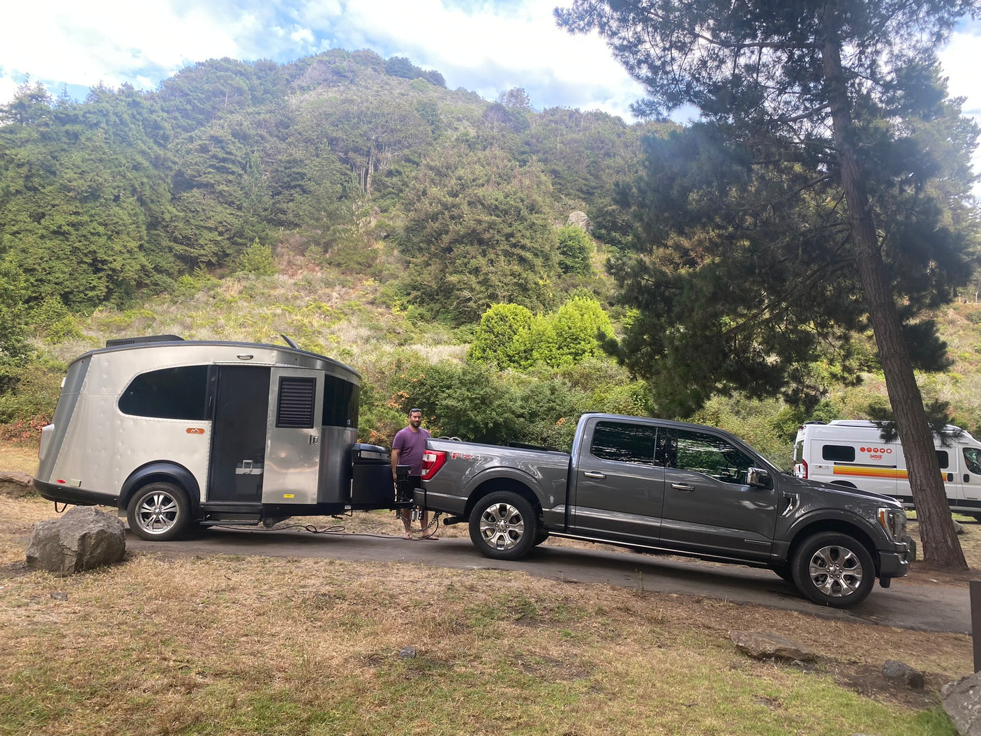 A man stands with his truck and a unique RV rental travel trailer at a beautiful forested campsite, near another camper van.