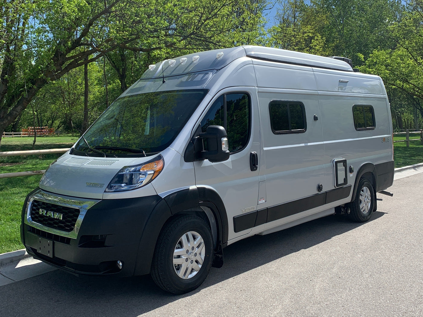 A light grey Winnebago Solis camper van is parked on asphalt, surrounded by green trees, perfect for an RV rental.