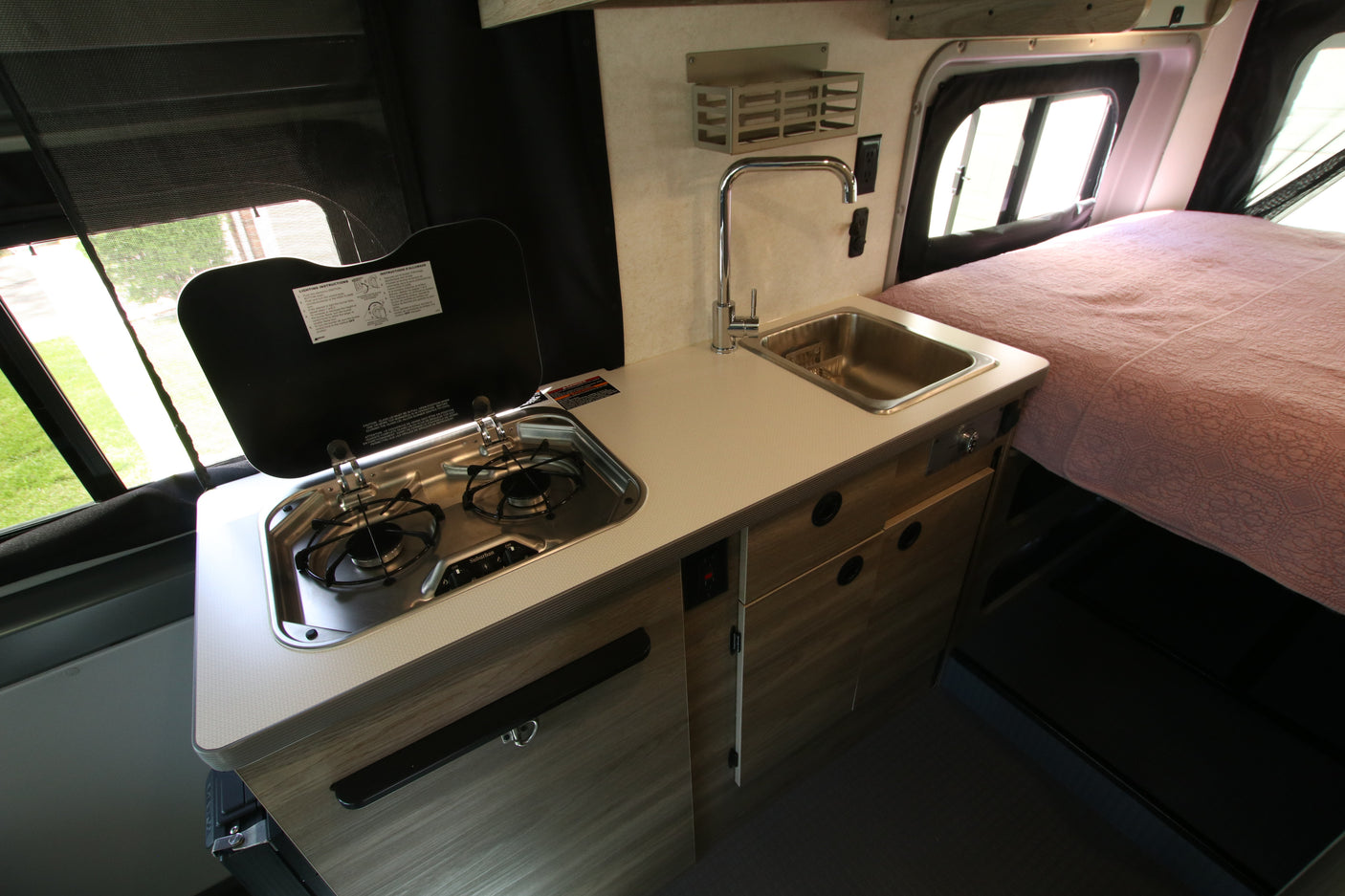 This RV rental camper van features a compact kitchen with a two-burner stove, sink, and a cozy bed area.