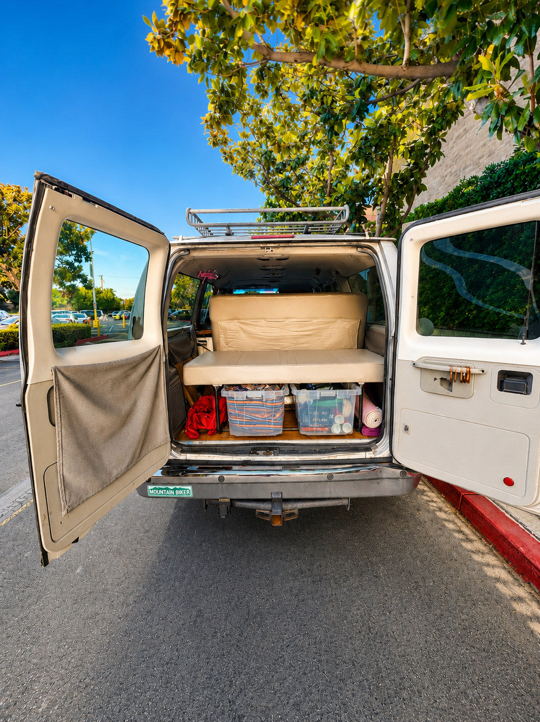 A custom white camper van with its rear doors open, showing a bed, storage bins, and a roof rack. Great RV rental for trips!