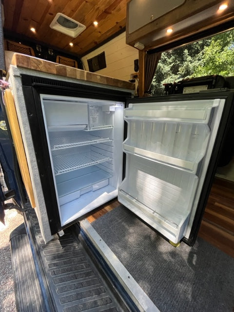 Inside this RV rental: open compact refrigerator showing freezer and fridge shelves. Perfect camper van kitchen setup.