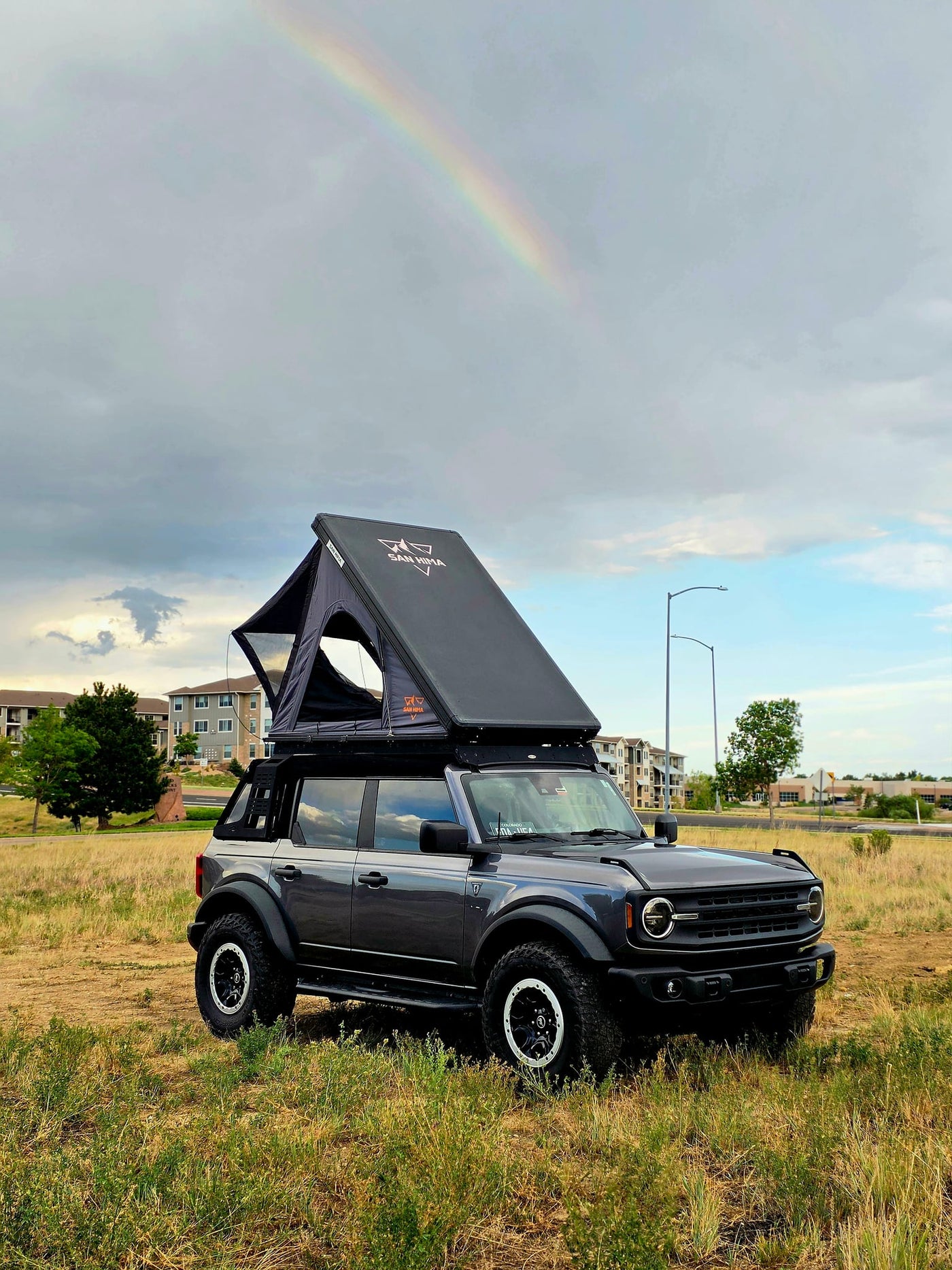 parked by hills and a highway overpass.|Gray Ford Bronco with an open rooftop tent in a grassy field. An ideal RV rental for off-road camping and outdoor adventure.|A Ford Bronco with a rooftop tent — photo 3
