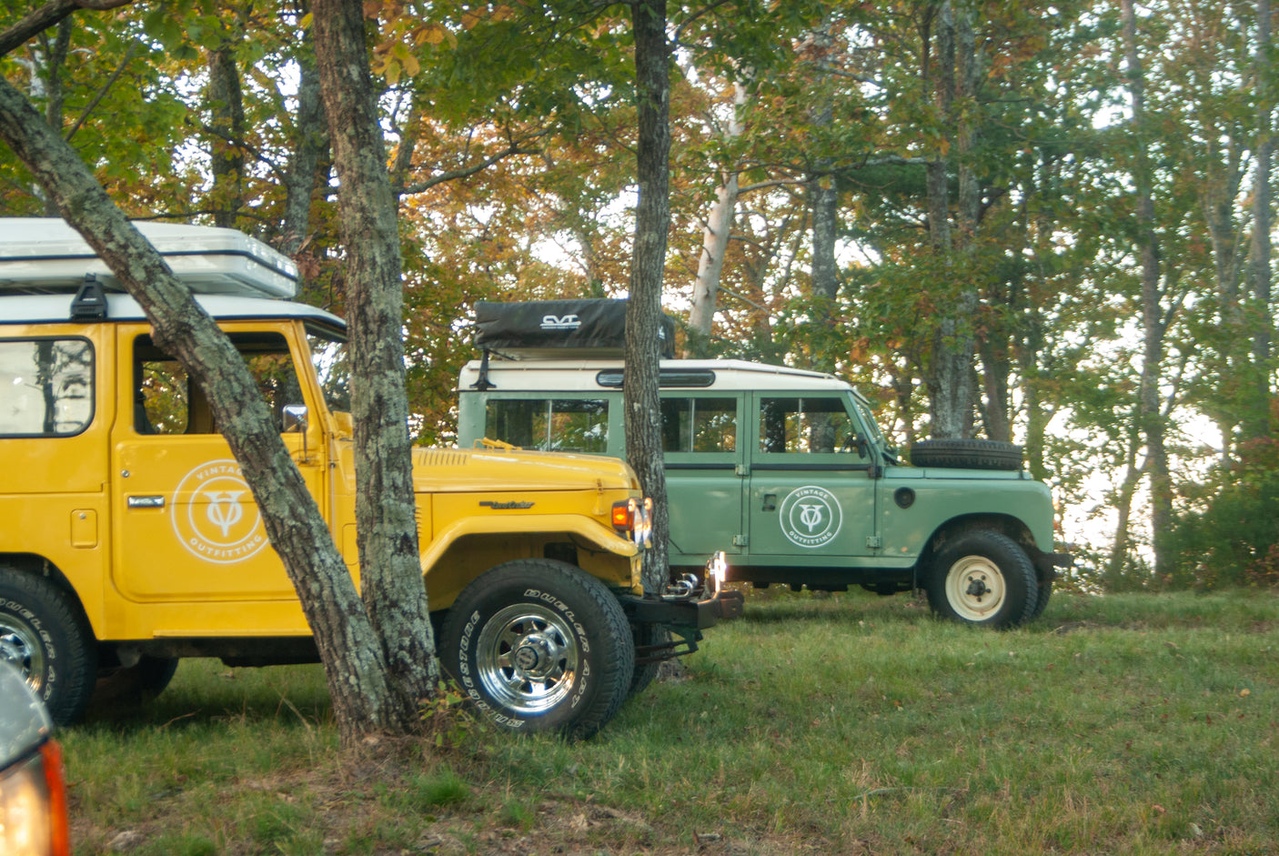 Two classic 4x4s, a yellow FJ40 & green Land Rover, parked for an RV rental adventure in the woods.