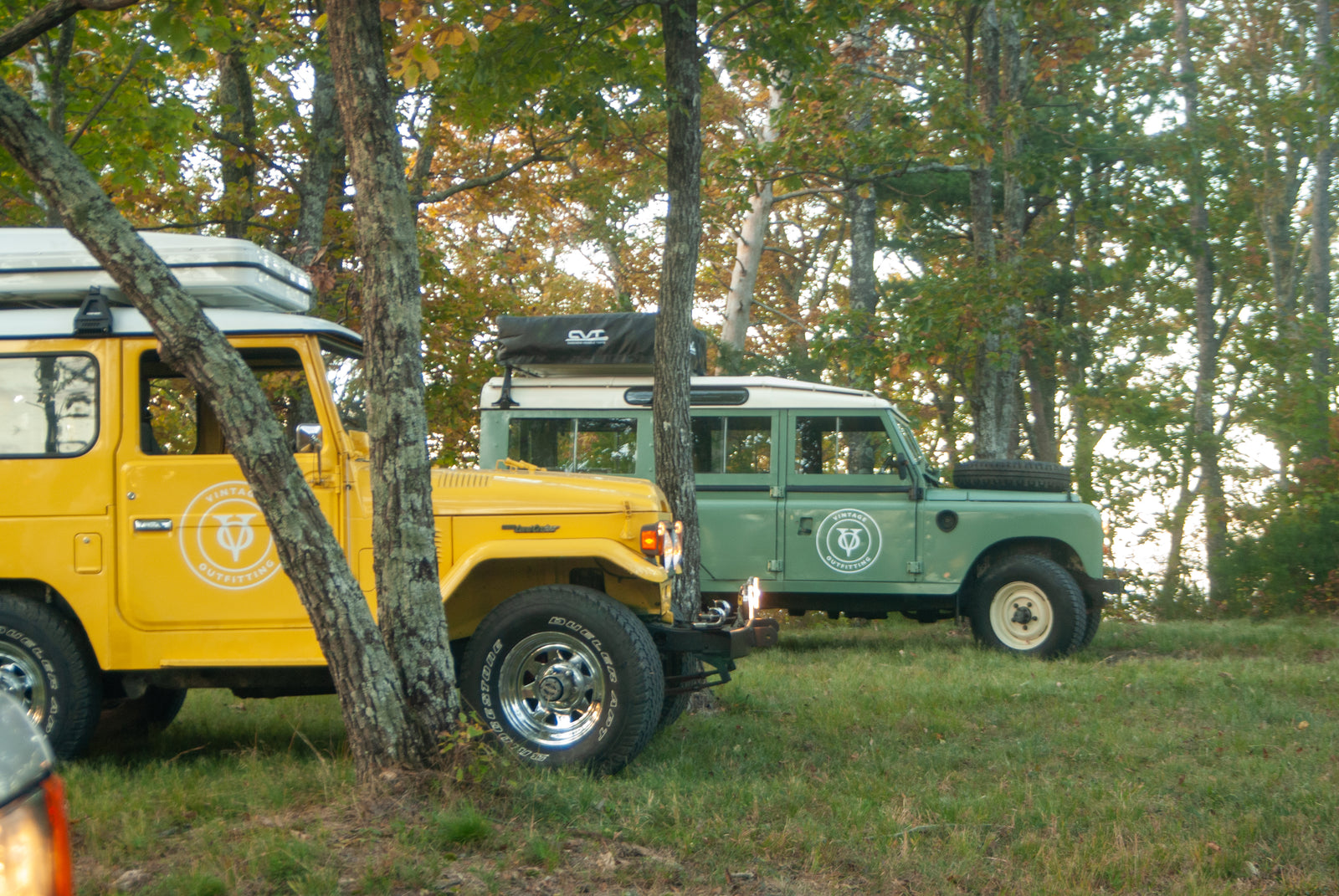 Two classic 4x4s, a yellow FJ40 & green Land Rover, parked for an RV rental adventure in the woods.