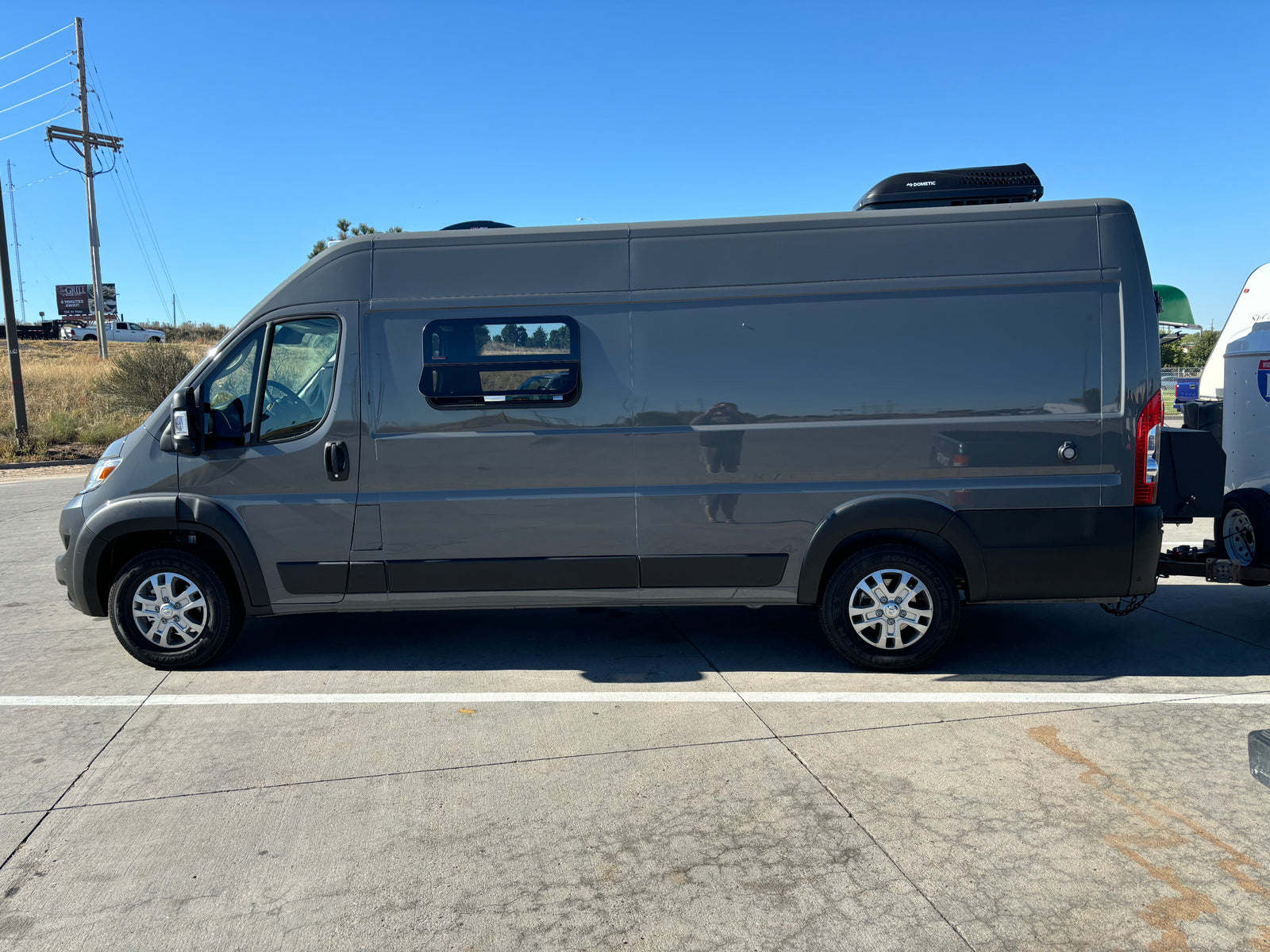 Grey RV rental camper van with a Dometic roof unit, parked on concrete, ready for travel and towing a trailer.