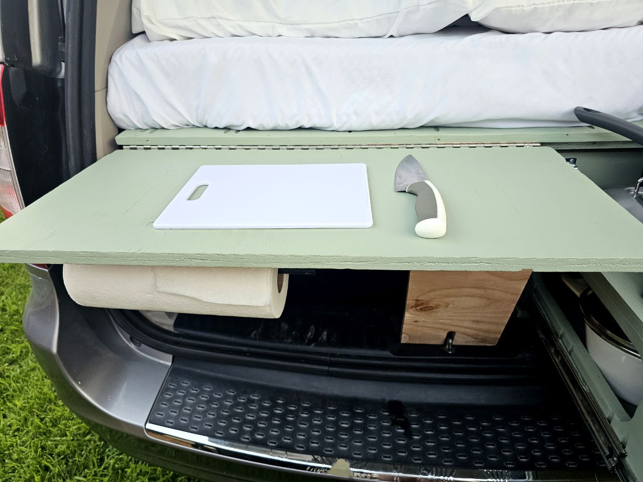 RV rental setup: Van's rear converts into an outdoor kitchen with a cutting board, knife, paper towels, and a bed above.