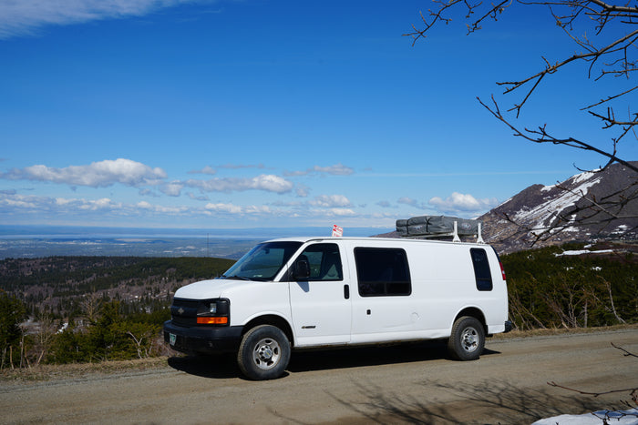 White rv rental van with rooftop tent parked on a mountain road, overlooking a vast forest and distant water under blue sky.