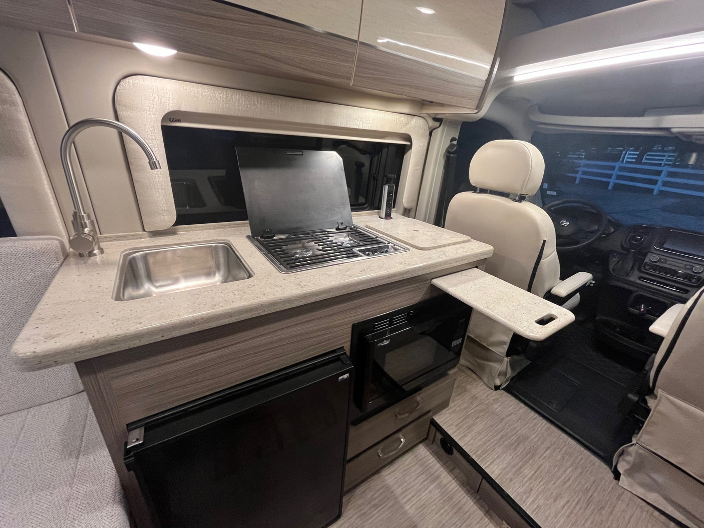 Compact RV rental interior: kitchen with sink, stovetop, microwave, fridge. Swivel front seats, extended table shown.