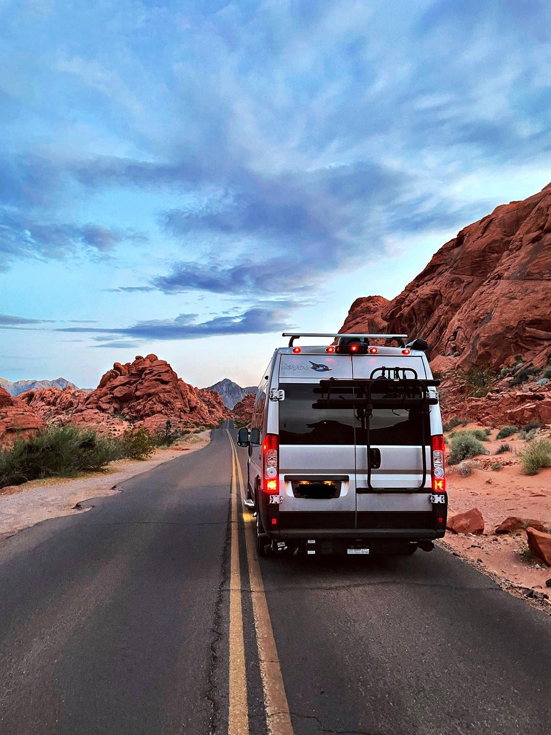 Back view of an RV rental driving on a desert road amidst towering red rocks under a vast, cloudy sky.