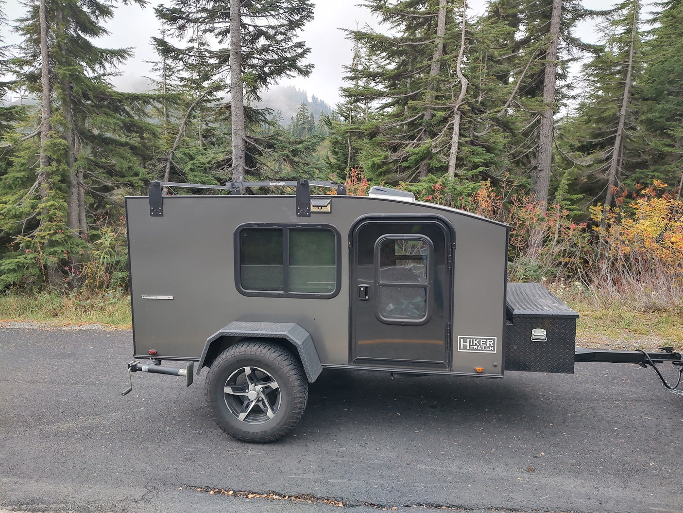 This gray Hiker Trailer RV rental is parked on asphalt, framed by tall evergreen trees and autumn leaves.